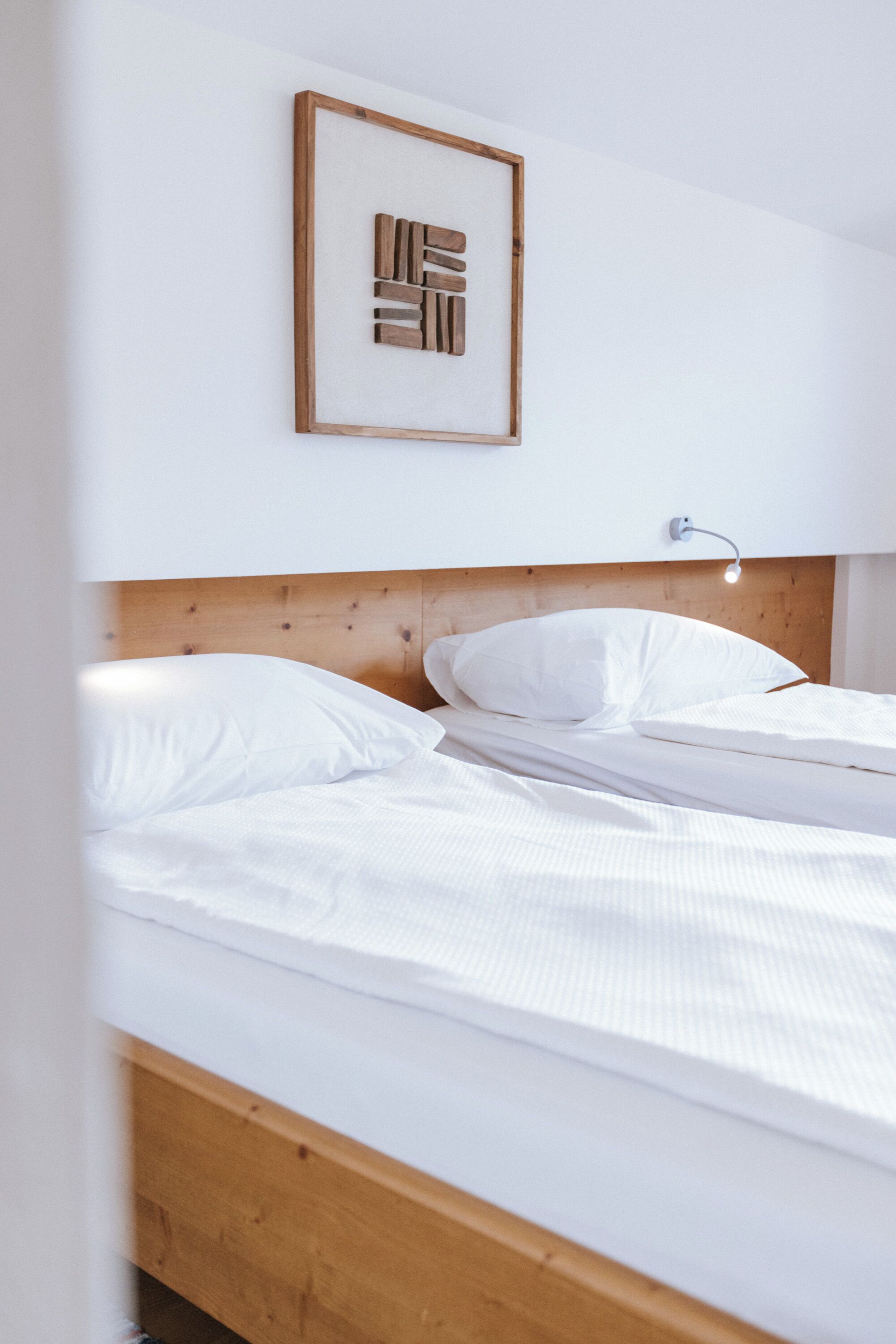 A bed with white linens, a wooden headboard, a wall-mounted light, and a framed abstract artwork.