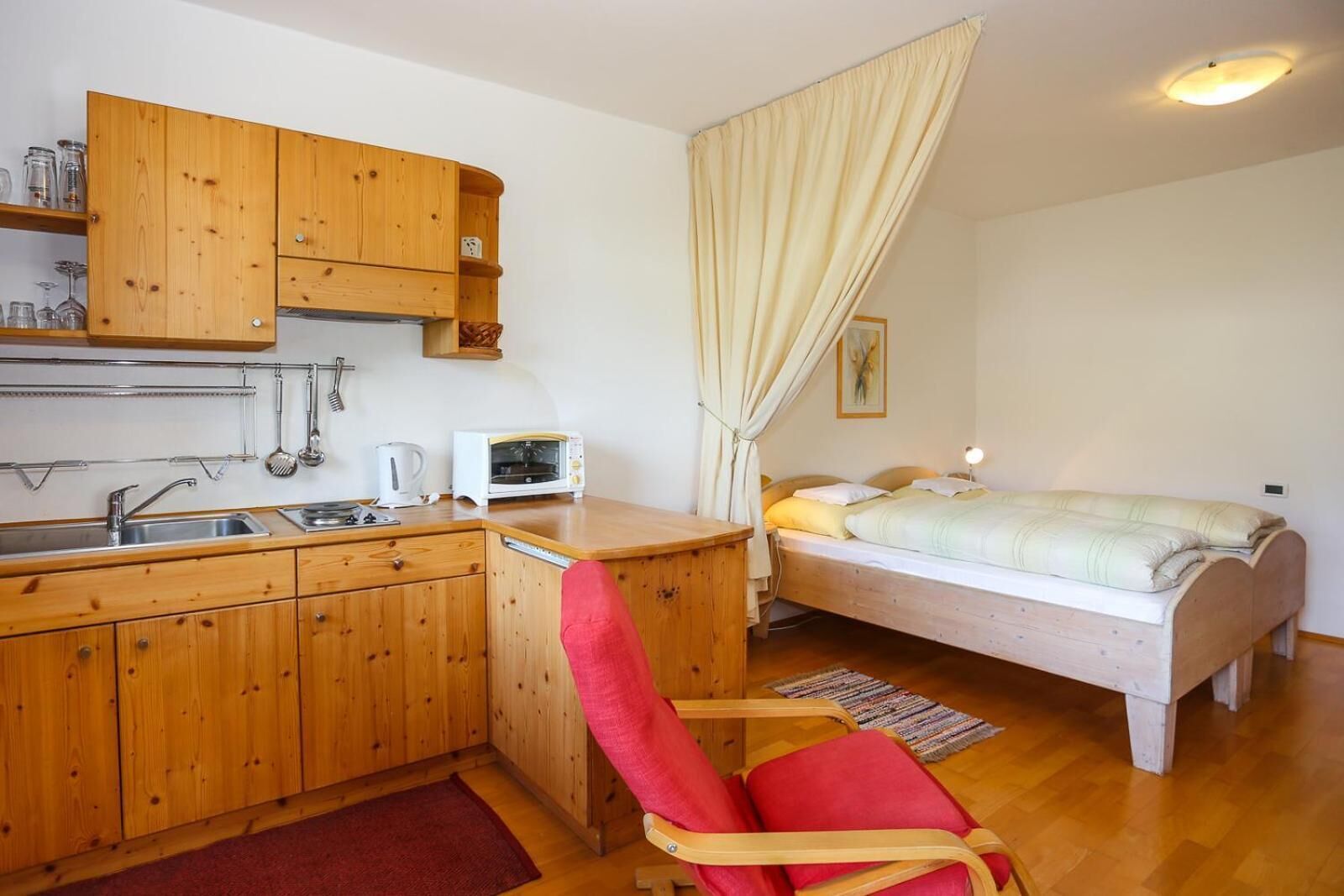 Una habitación compacta con kitchenette, una cama y una silla roja.