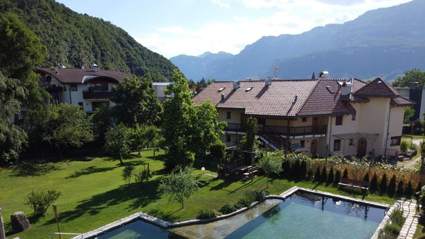 A swimming pool with a wooden deck, surrounded by a well-maintained lawn and trees, with a mountain backdrop. - Bio-Hotel & Residence Kaufmann (Ora)