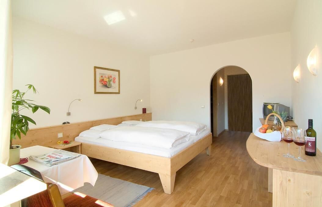 A bedroom with a wooden bed, a desk with a chair, a potted plant, a framed picture on the wall, and a small table with wine glasses and a basket of fruit.