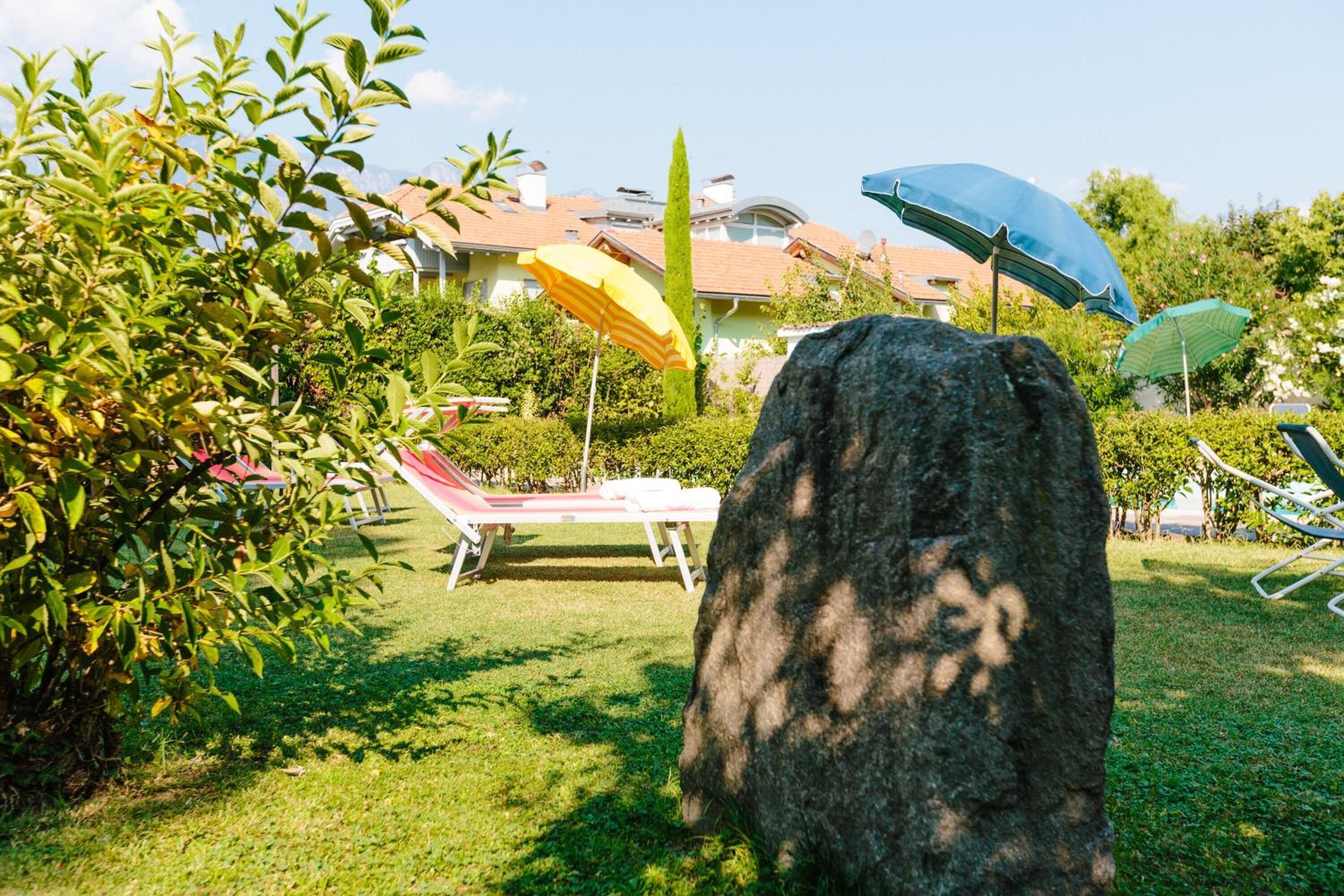 Un jardín con una roca grande, una zona de piscina con sillones y sombrillas, y una casa al fondo.