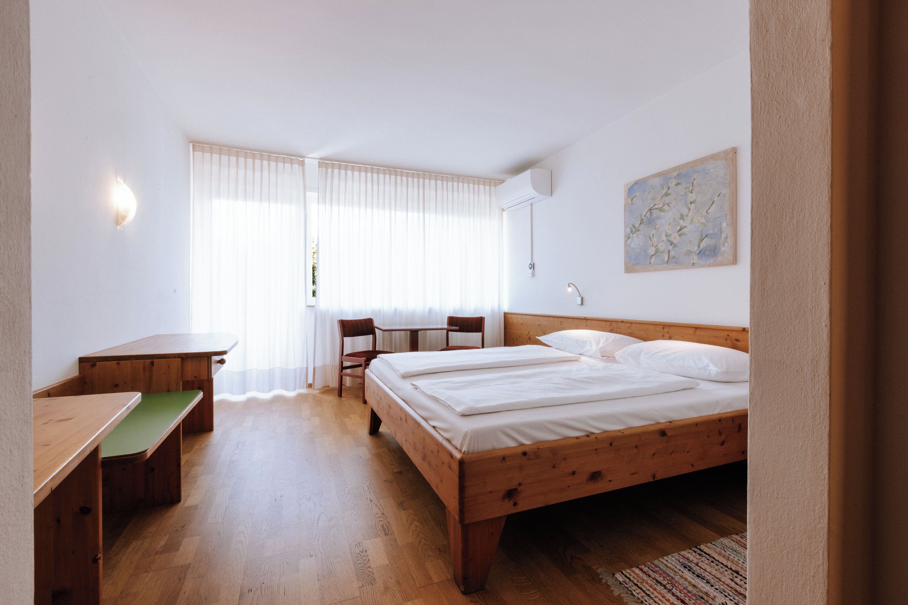 A bedroom with a wooden bed, a desk with a green bench, a chair, and a window with curtains.
