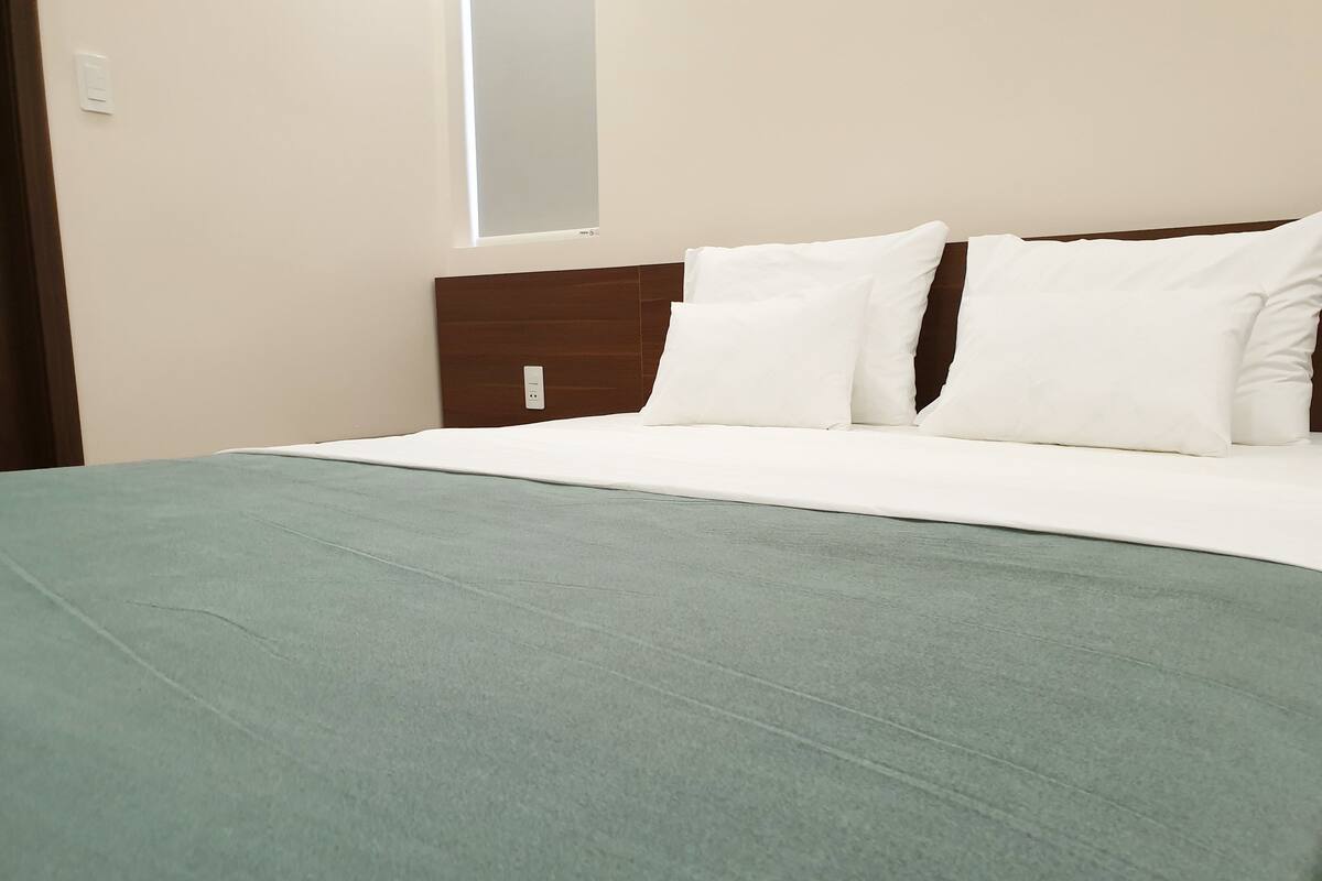 A neatly made bed with a green bedspread and white pillows, a wooden headboard, and a window with blinds.