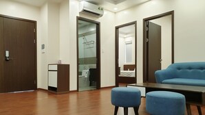 A modern hotel room with wooden flooring, a blue sofa, two matching ottomans, a desk, and a glass-enclosed shower area. - Pure Stay (Hai Phong)