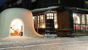 Un edificio con una gran estructura cubierta de nieve al frente, fachada de madera y un interior iluminado visible a través de las ventanas.