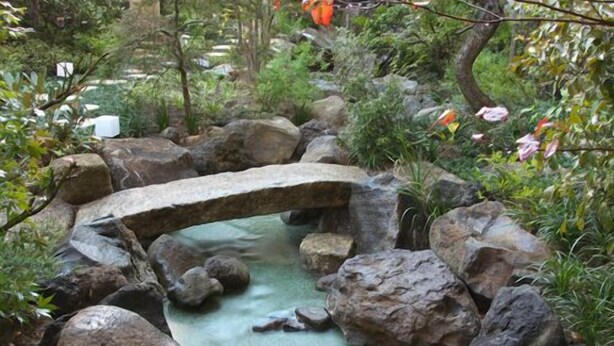 Un jardín con un puente de piedra sobre un arroyo, rodeado de rocas y follaje.