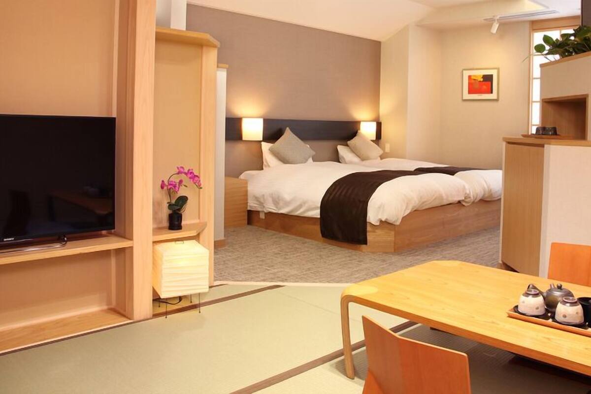 A hotel room with a bed, bedside table, TV, and a dining area with chairs and a table.