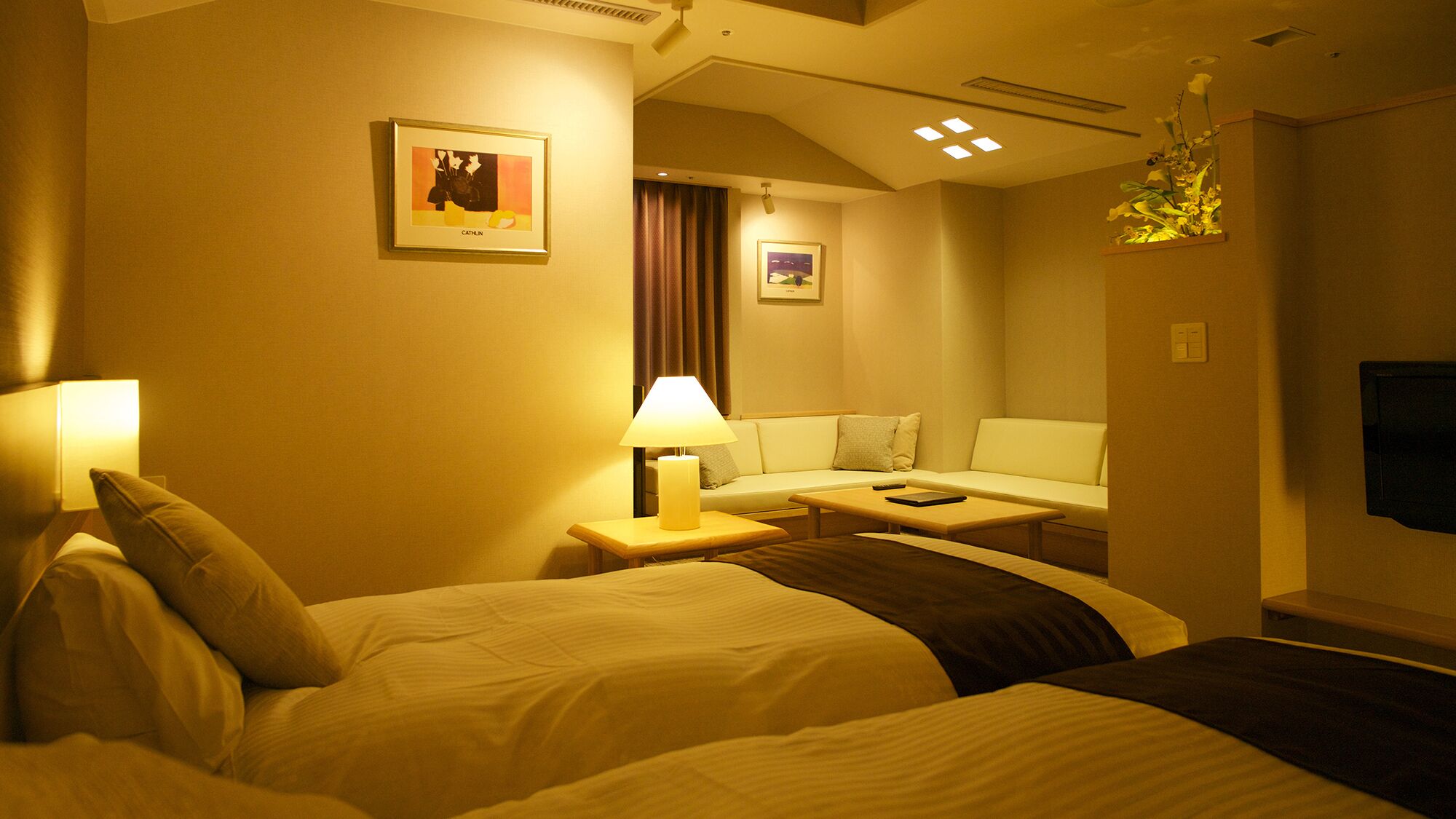 A hotel room with two beds, a sofa, a table, a lamp, and artwork on the walls.