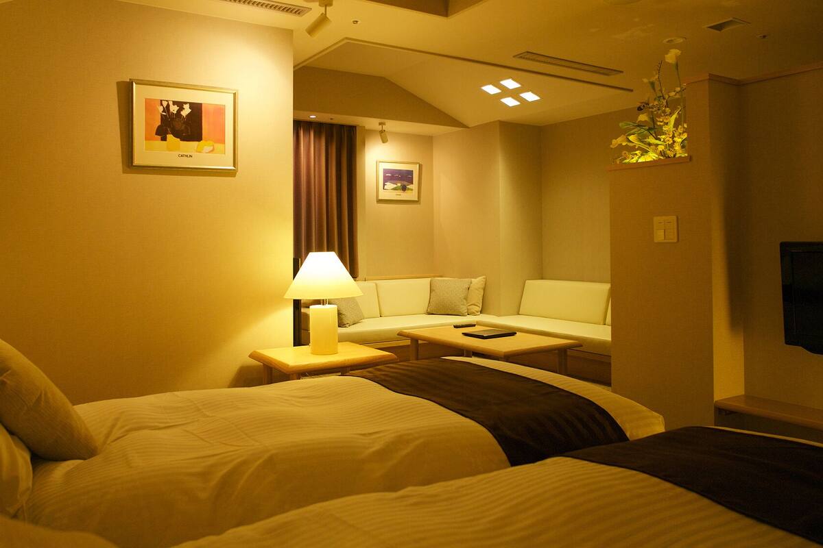 A hotel room with two beds, a sofa, a table, a lamp, and artwork on the walls.