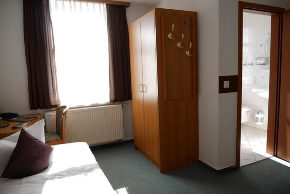 Habitación de hotel con cama, armario de madera, escritorio con silla y vista al baño a través de una puerta abierta.