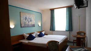 Ein Hotelzimmer mit Bett, Schreibtisch, Stuhl, einem Bild an der Wand und einem Vorhangfenster.