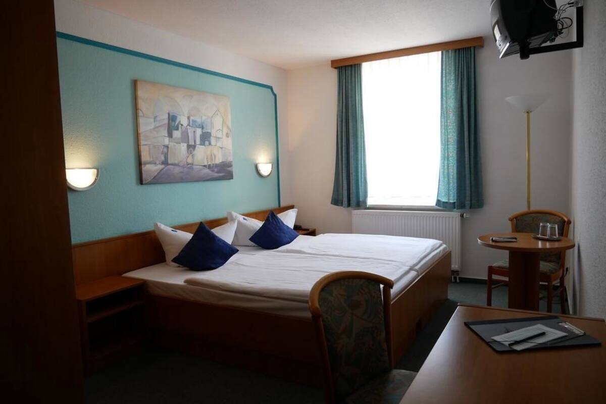 Et hotelværelse med en seng, et skrivebord, en stol, et billede på væggen og et vindue med gardiner.