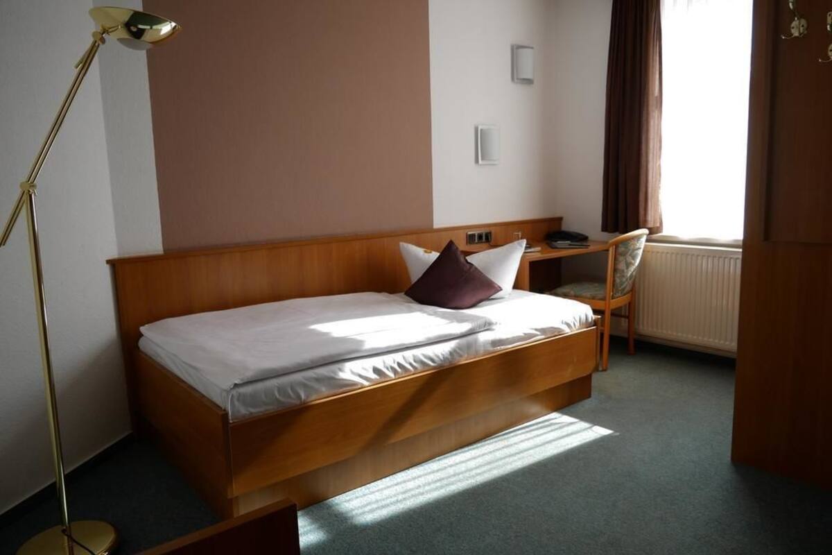 En enkeltseng med træsengegavl, et natbord, en stol og en gulvlampe i et rum med et vindue og en radiator.