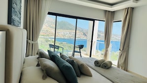 Une chambre avec une grande fenêtre donnant sur un paysage pittoresque, avec une étendue d’eau et des collines en toile de fond. On y trouve un lit avec des oreillers et une serviette enroulée posée dessus.