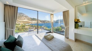 Une chambre moderne avec un grand lit, deux fauteuils, une table de chevet et une vue sur la mer et les montagnes.