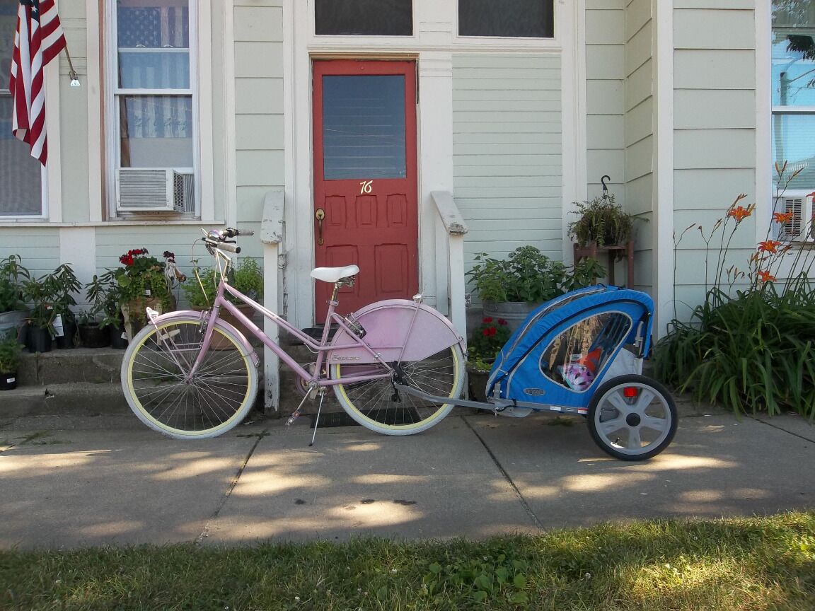 Uma bicicleta rosa com um reboque, estacionada em frente a uma casa com porta vermelha e o número 76.