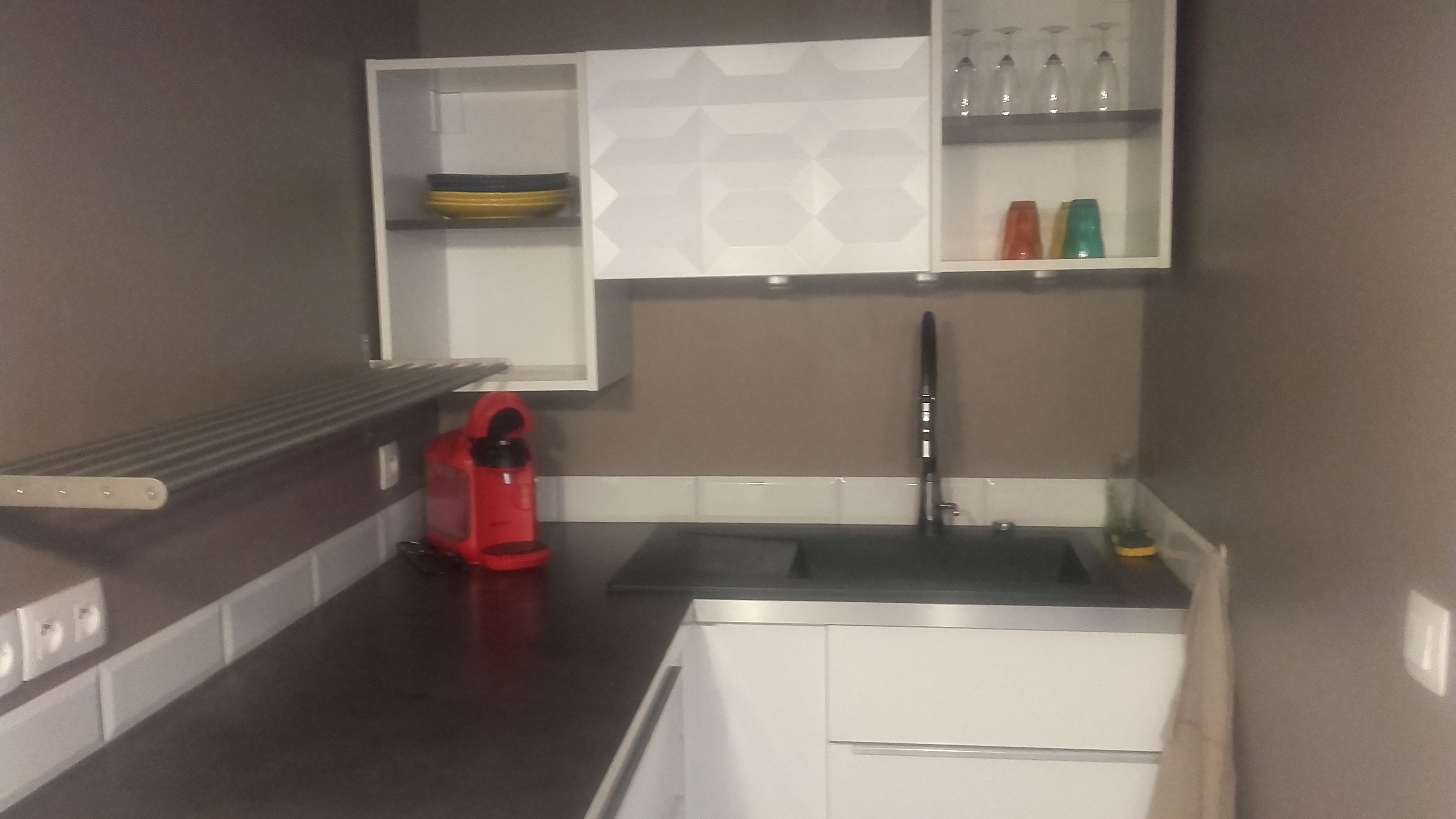 Cocina compacta con pileta integrada, una tetera roja y una estantería con vasos y botellas.