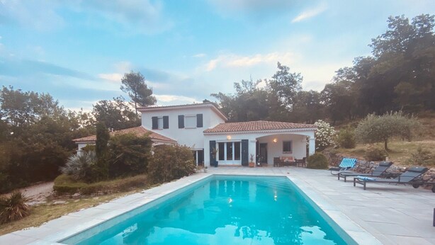 Una piscina con borde de baldosas, una casa con techo de tejas y un área de patio con sillas de descanso.