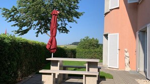 Una mesa de picnic con un paraguas rojo, una estatua y un edificio con contraventanas blancas.