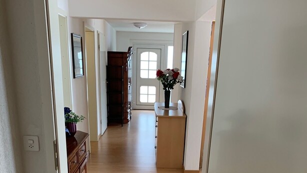 Un pasillo con suelo de madera, una puerta con manija de latón y un jarrón con flores sobre una mesita.