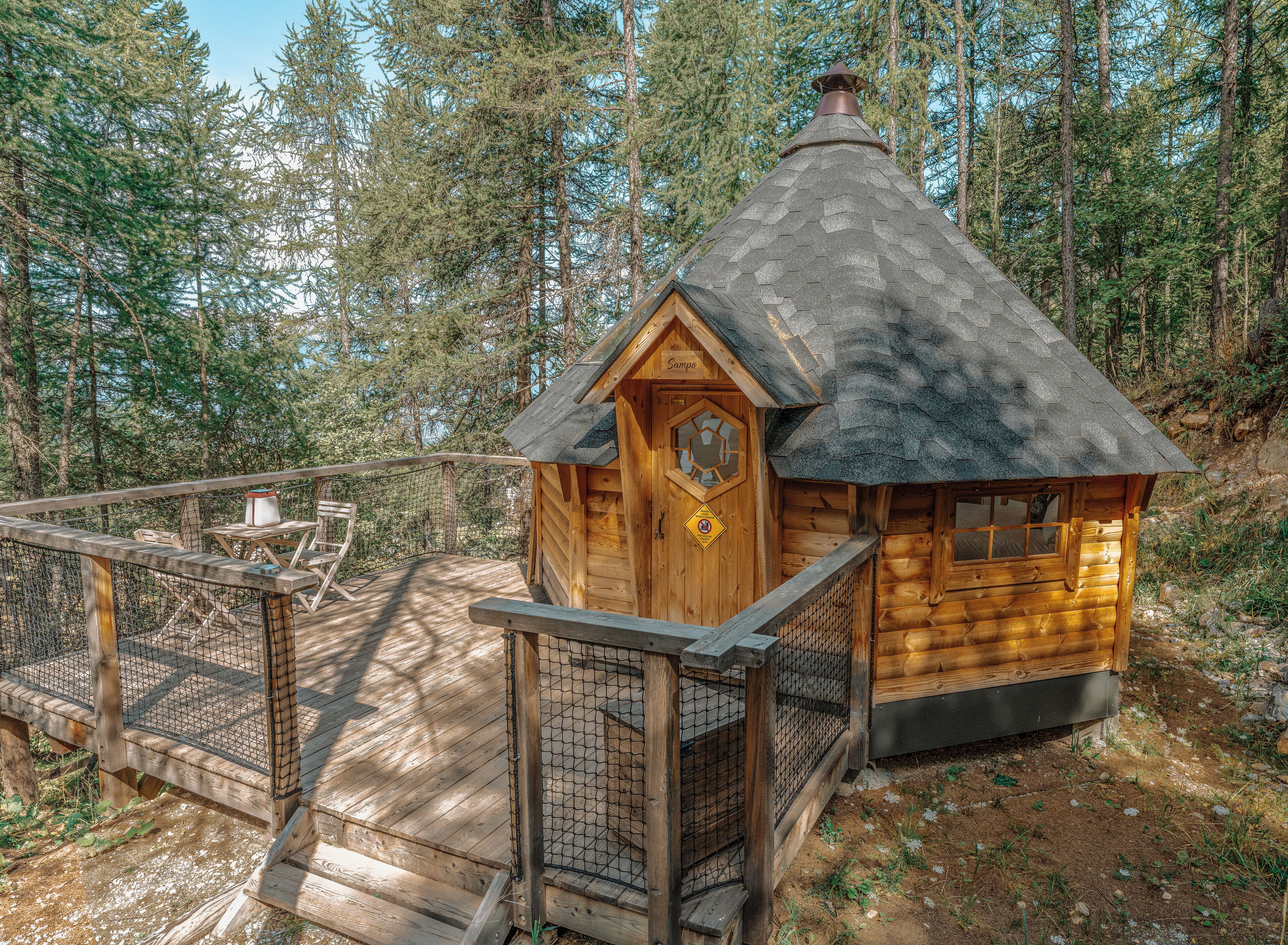 A wooden cabin with a thatched roof, surrounded by a wooden deck and a metal fence, set in a forested area.