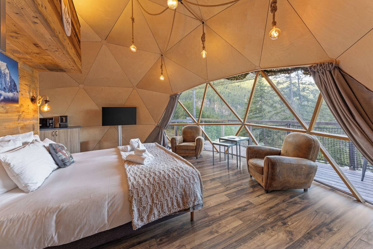 Quarto espaçoso com uma cama grande, duas poltronas, uma mesa pequena e uma TV. O quarto tem piso de madeira, uma janela ampla com vista para árvores e teto com luminárias pendentes.