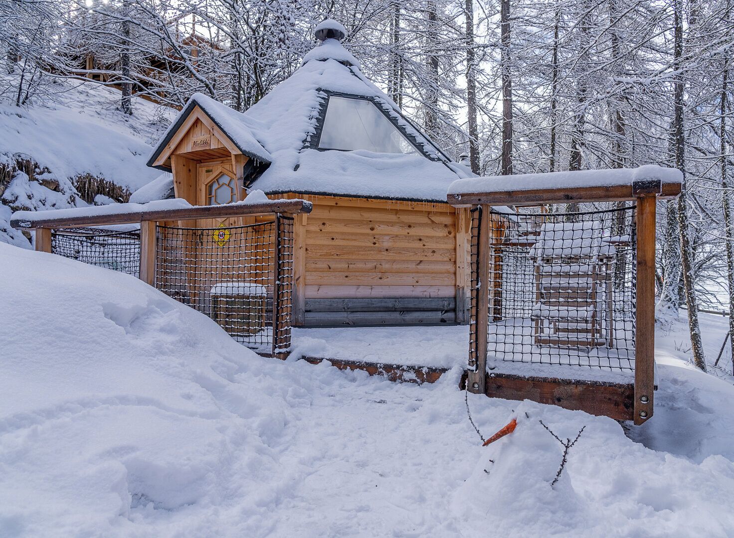 A wooden cabin with a snow-covered roof, surrounded by a wooden fence and snow-covered ground.