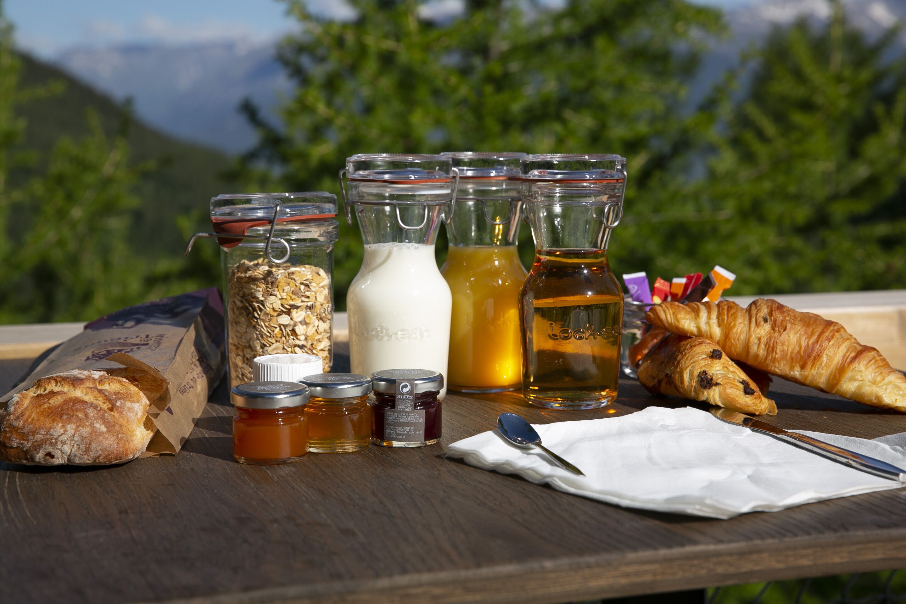 A table with food items, including bread, jars of honey, and a bottle of tea, set outdoors with a mountain view in the background.