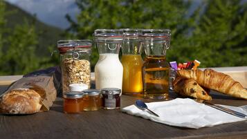 A table with food items, including bread, jars of honey, and a bottle of tea, set outdoors with a mountain view in the background.