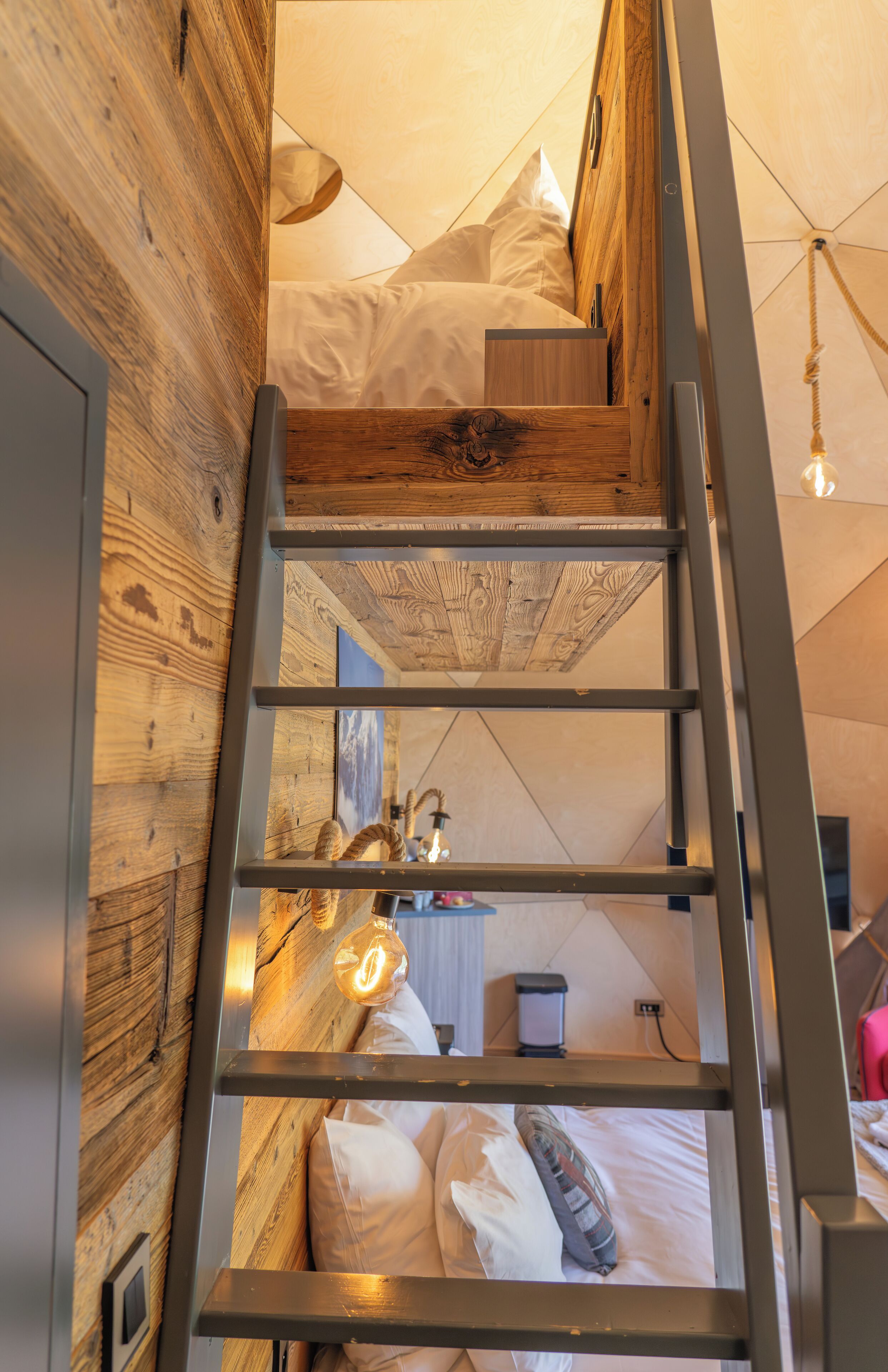 A loft-style bedroom with a wooden floor, a ladder leading to a bunk bed, and a hanging light fixture.