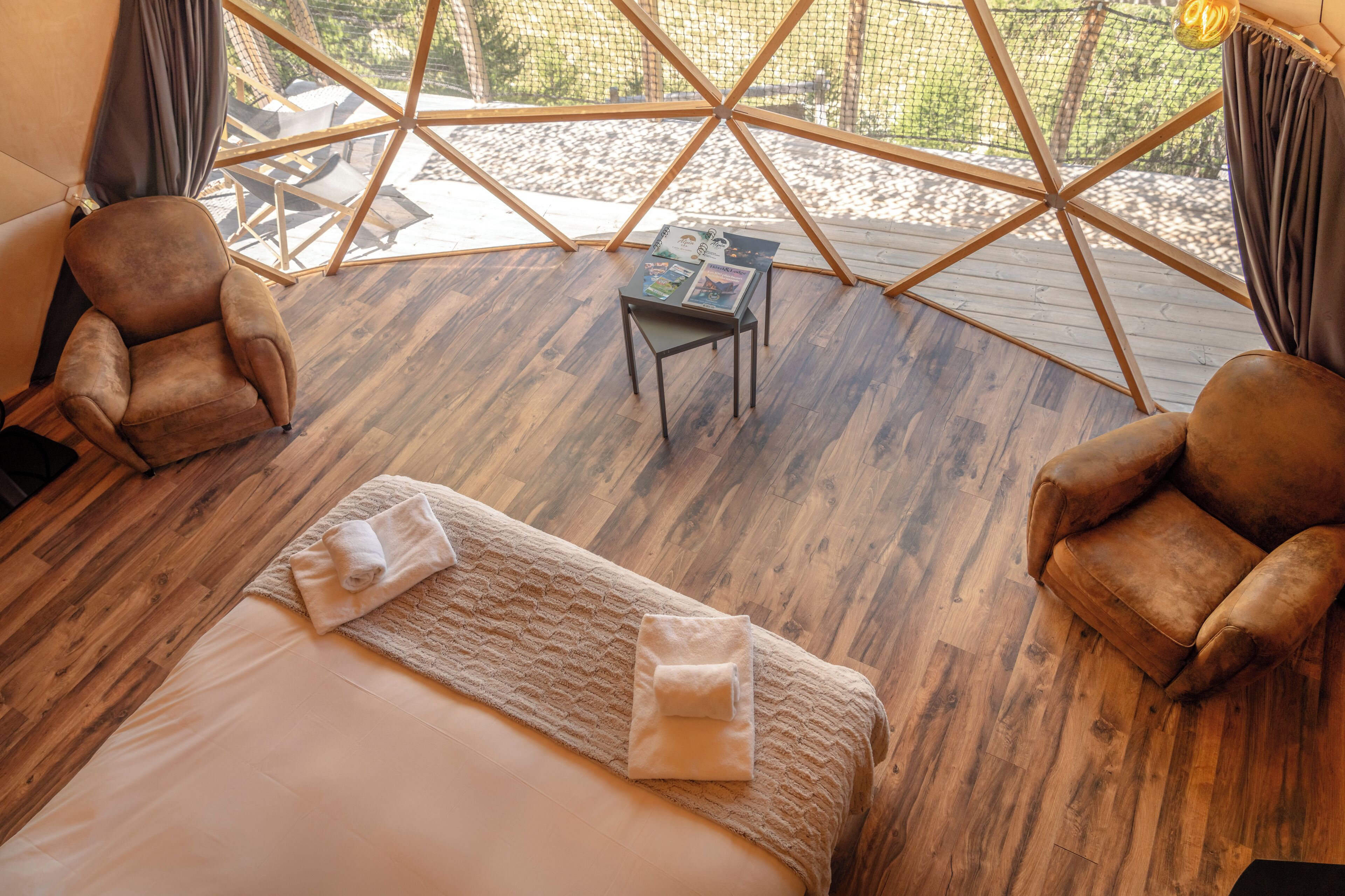 A cozy room with a wooden floor, a bed with a white comforter, two leather armchairs, and a small table with magazines.