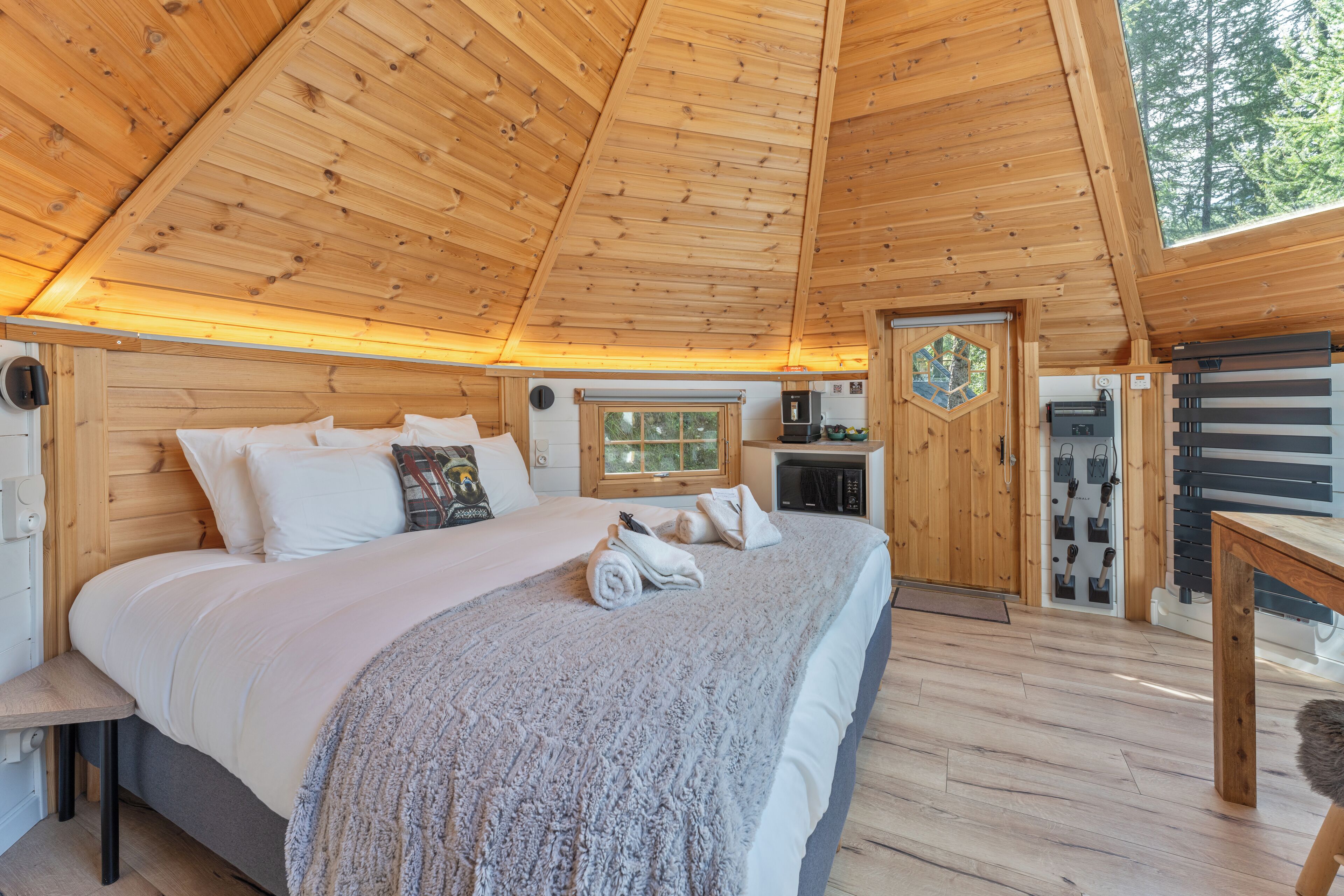 A wooden cabin bedroom with a bed, pillows, a blanket, a small table, and a window.