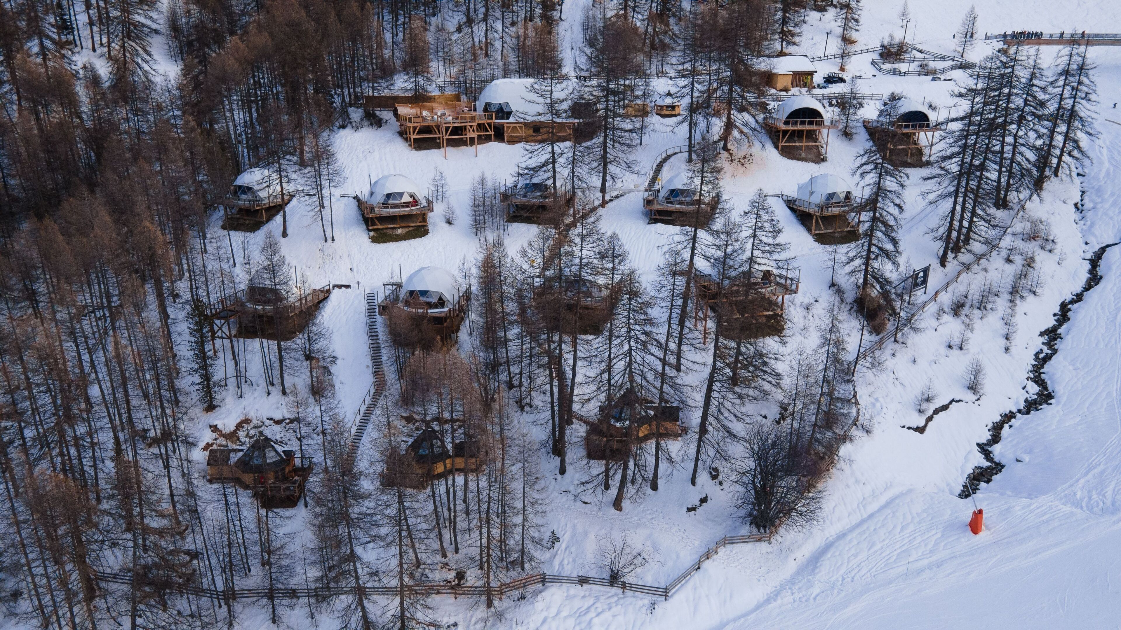 Vue aérienne d’un chalet enneigé, avec plusieurs cabanes en forme de dôme, des passerelles en bois et une zone boisée recouverte de neige.