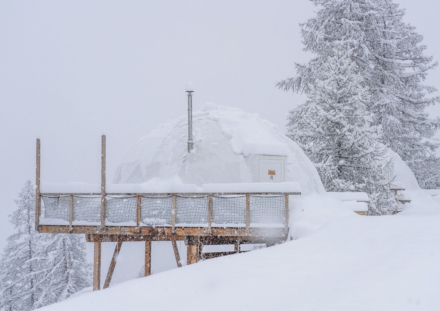 Een besneeuwde buitenruimte met een houten constructie, mogelijk een terras of platform, en een klein gebouw met een deur en een bordje.