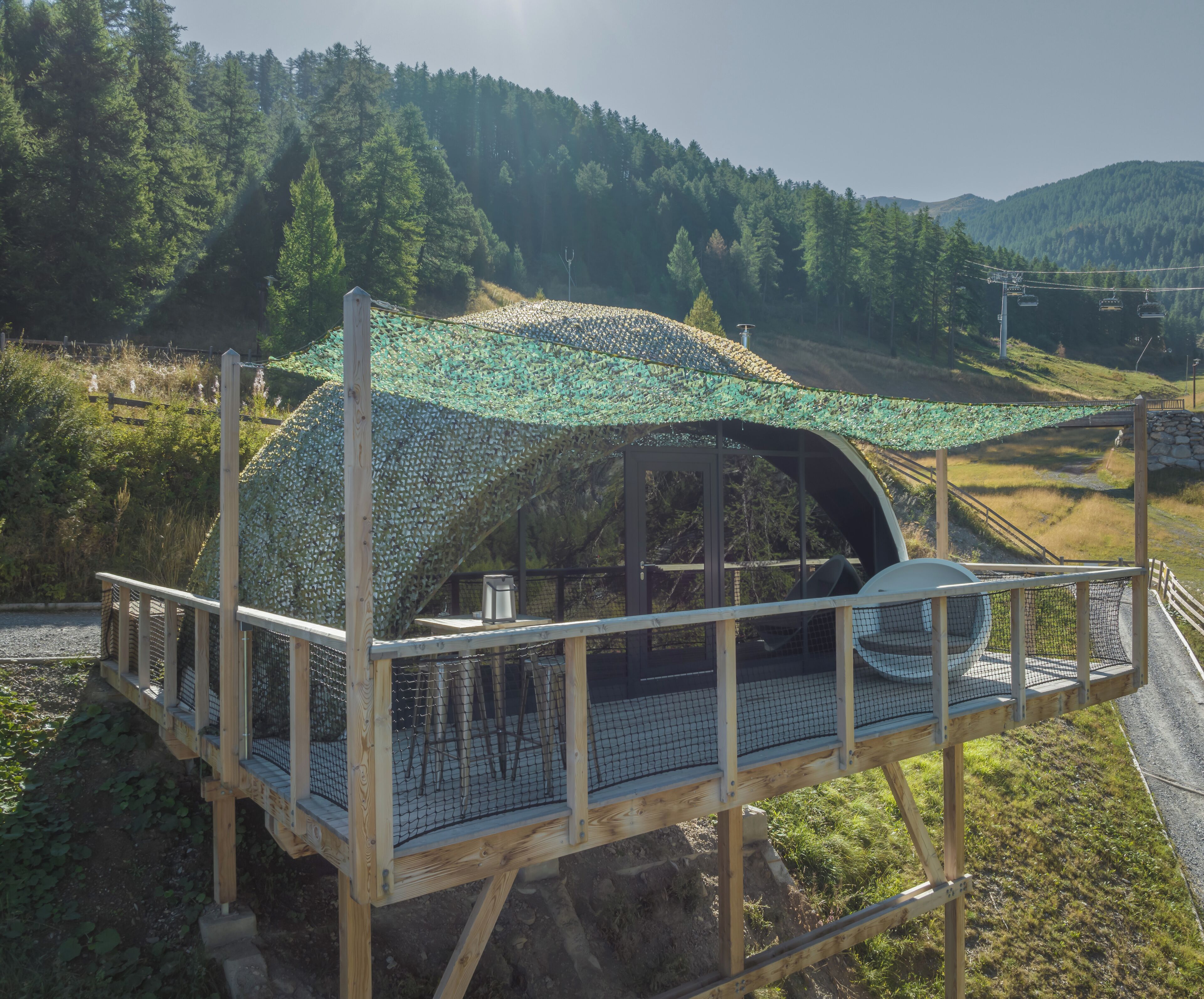 Una estructura de madera con techo curvo y baranda de metal, rodeada por un bosque en la ladera de una colina.