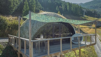 Una estructura de madera con techo curvo y baranda de metal, rodeada por un bosque en la ladera de una colina.