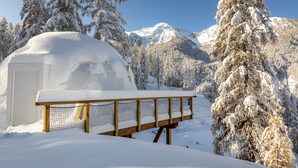 Una iglú cubierta de nieve con un banco de madera al frente, rodeada de árboles cargados de nieve y montañas.
