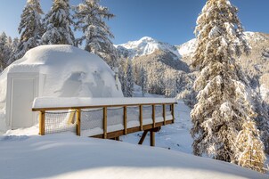 Un igloo recouvert de neige, avec un banc en bois devant, entouré d’arbres et de montagnes chargés de neige.