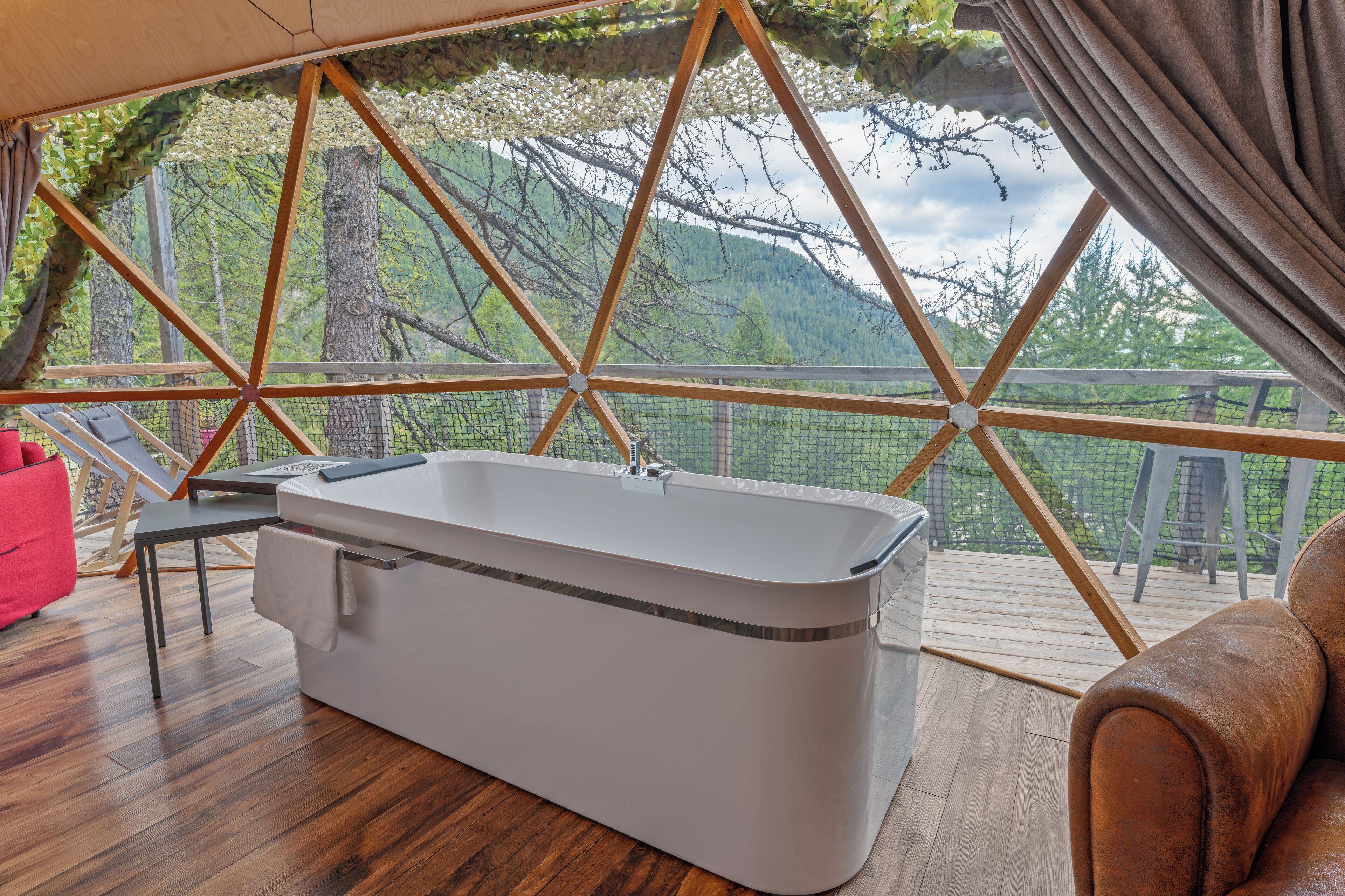 A modern bathroom with a bathtub, wooden flooring, and a large window offering a view of trees and hills.