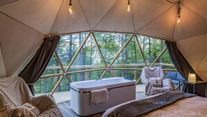 Una habitación acogedora tipo tienda de campaña, con una cama grande, un rincón de descanso con una silla y una mesa, una bañera y vistas a un bosque a través de un diseño de ventana geométrico.