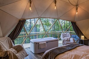 Une pièce accueillante, aménagée comme une tente, avec un grand lit, un coin salon comprenant une chaise et une table, une baignoire et une vue sur une forêt, grâce à une fenêtre au design géométrique.