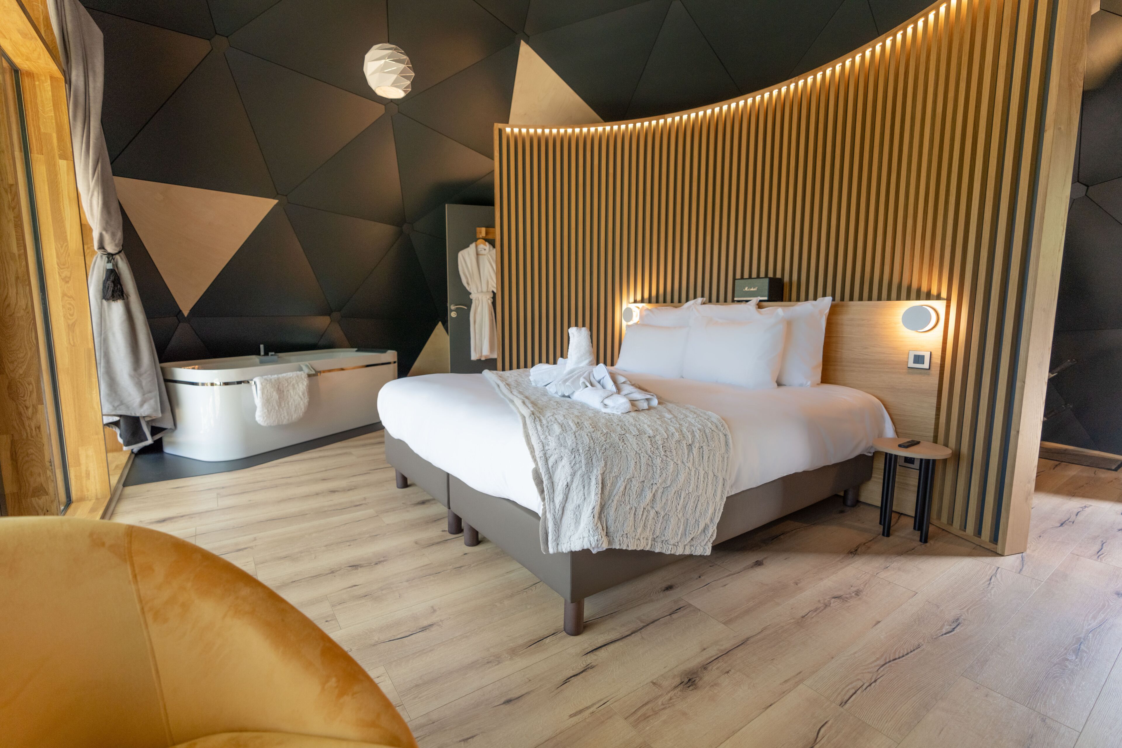 A modern hotel room with a large bed, a small bedside table, a wall-mounted light, and a wooden panel design.