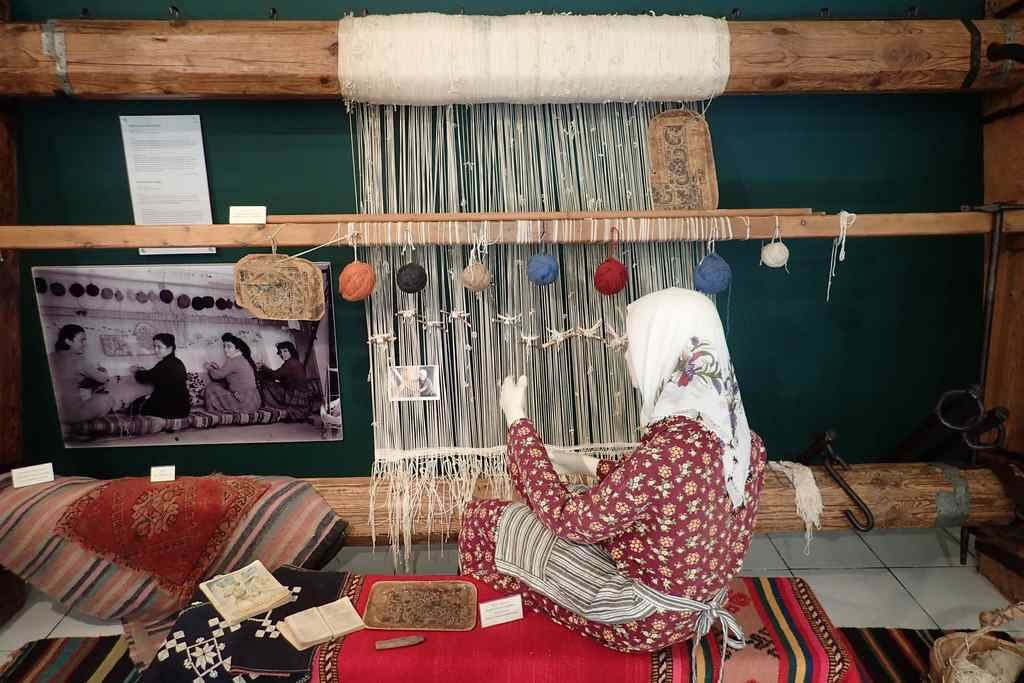 A person weaving on a loom with a traditional setup, including a wooden frame and a green wall.