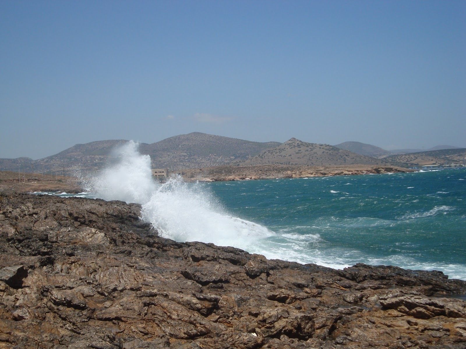 A rocky coastline with waves crashing against the shore, a clear sky, and distant mountains.