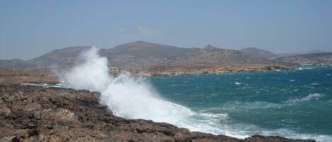 A rocky coastline with waves crashing against the shore, a clear sky, and distant mountains.