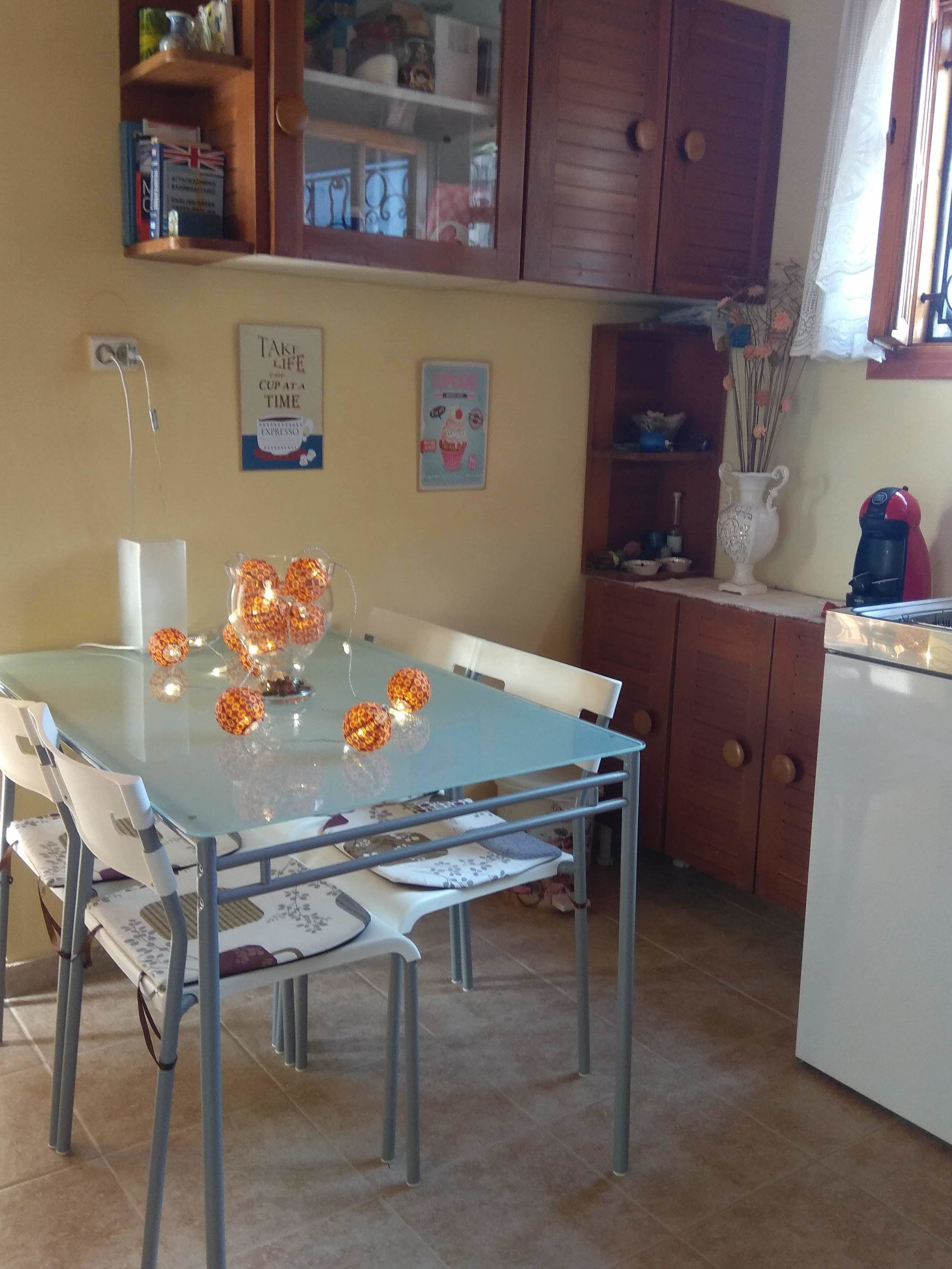 Een eetgedeelte met een glazen tafel, witte stoelen en decoratieve oranje bollen. Er is een houten kast met planken boven de tafel, en een kleine witte koelkast aan de rechterkant.