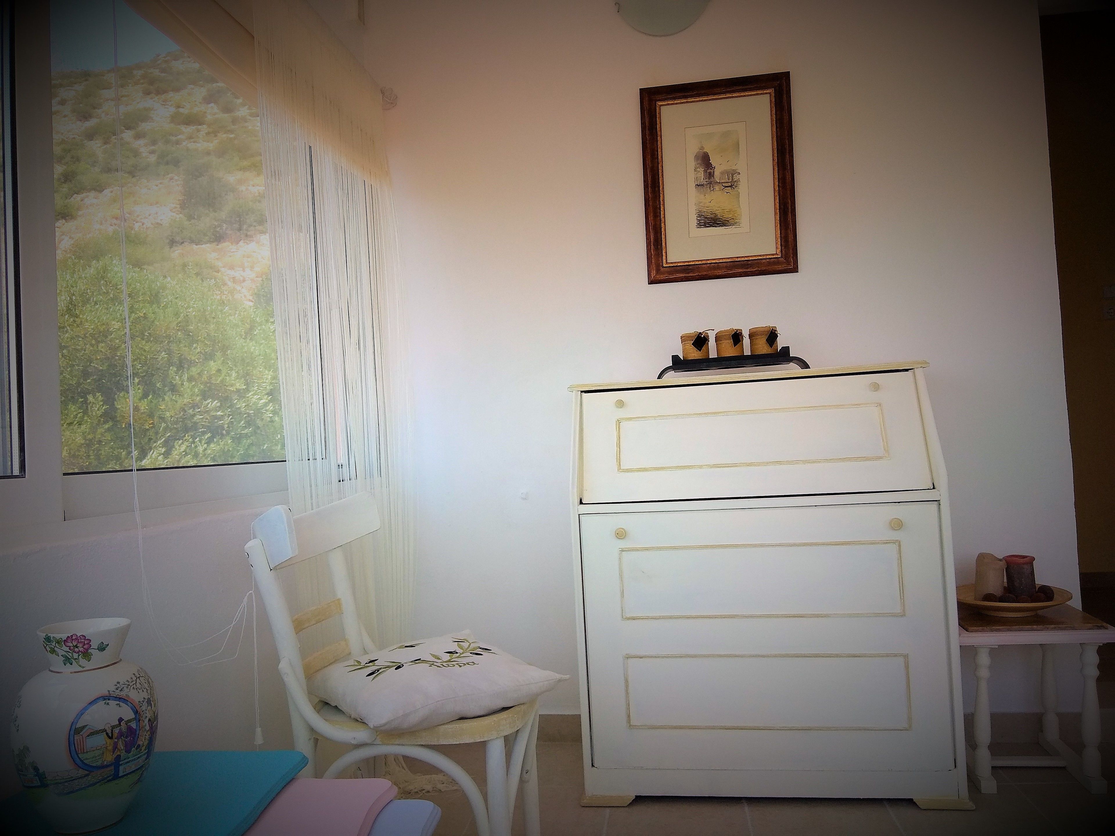 A room with a white dresser, a chair with a cushion, a small table with a bowl, and a framed picture on the wall.