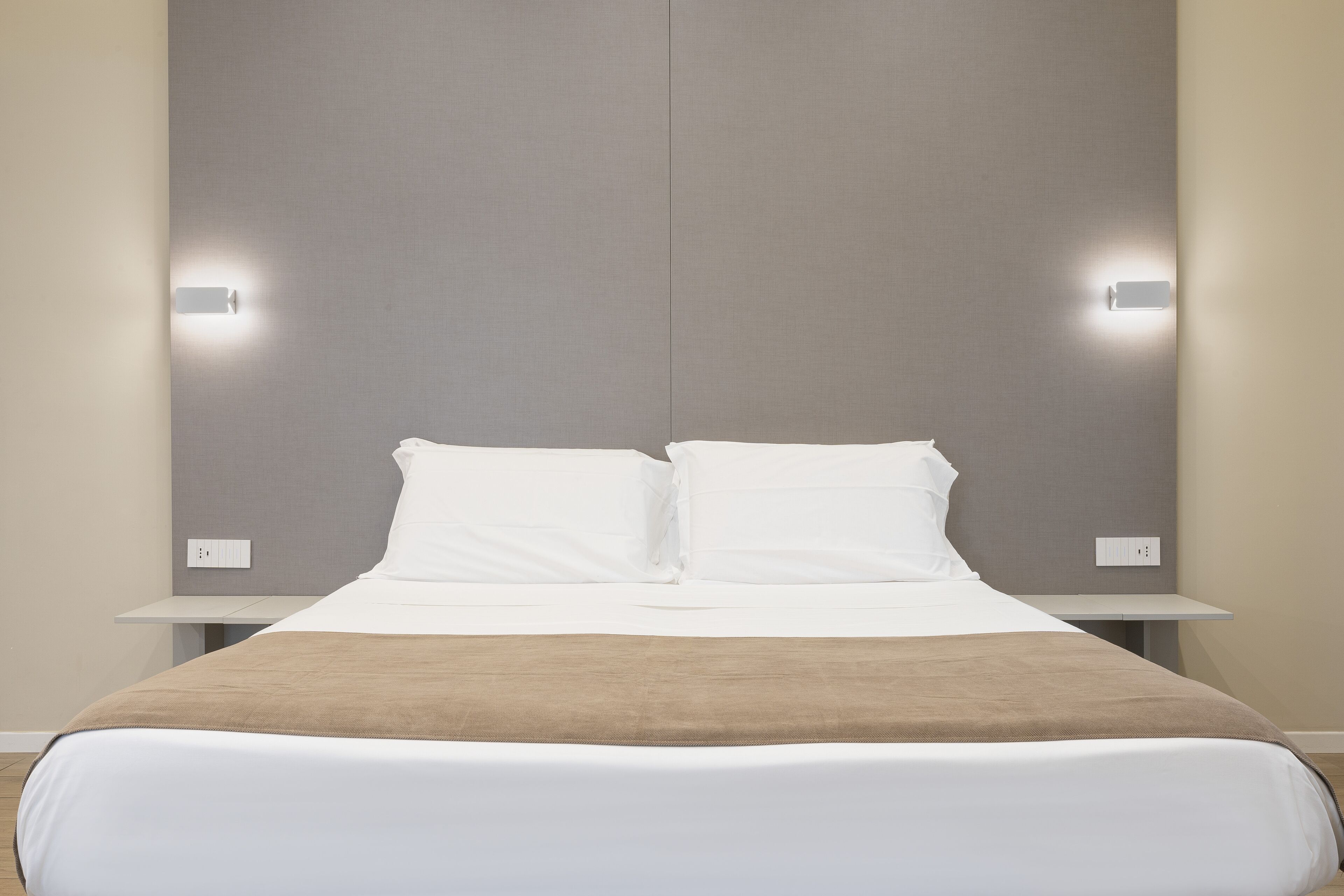 Een net opgemaakt bed met witte lakens en een bruine plaid, met aan beide zijden nachtkastjes met wandlampen.
