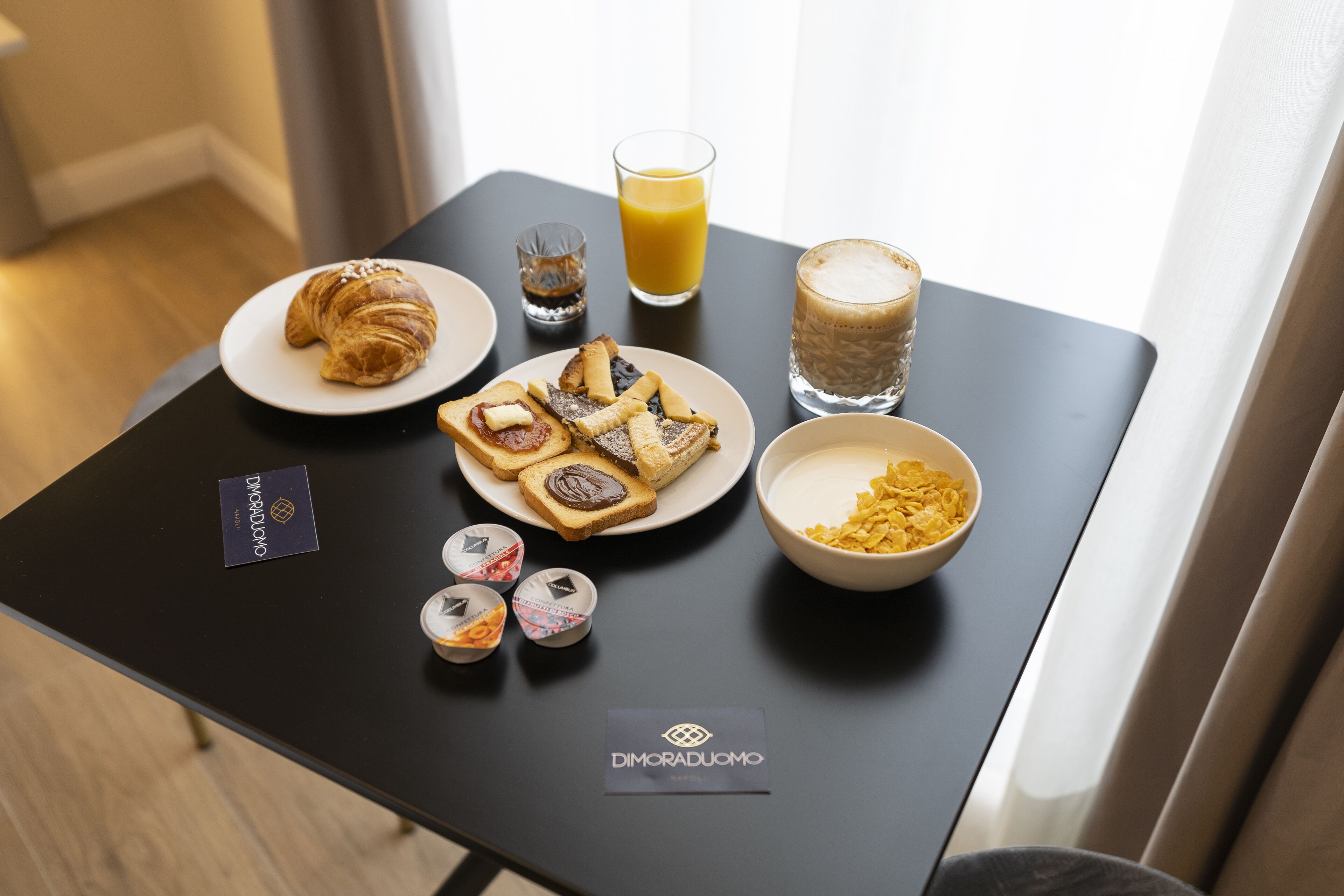 A breakfast table with croissants, toast with jam, a glass of orange juice, a glass of coffee, and a bowl of cereal.