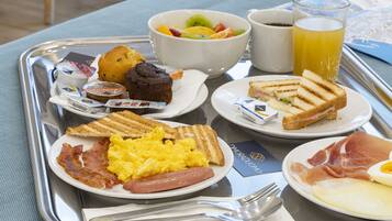 Un plateau de petit-déjeuner avec des œufs, du bacon, des saucisses, des toasts, des muffins et un saladier de fruits.